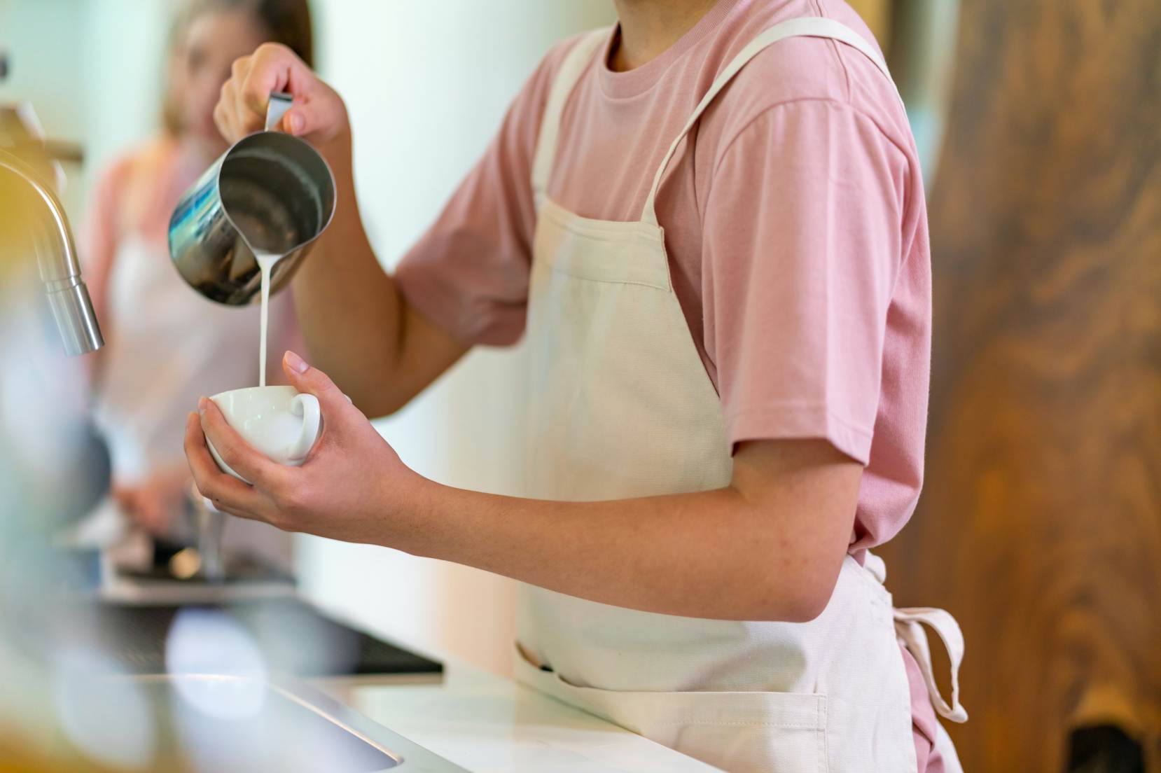 Donna che prepara un cappuccino. La donna indossa una maglietta rosa e un grembiule bianco